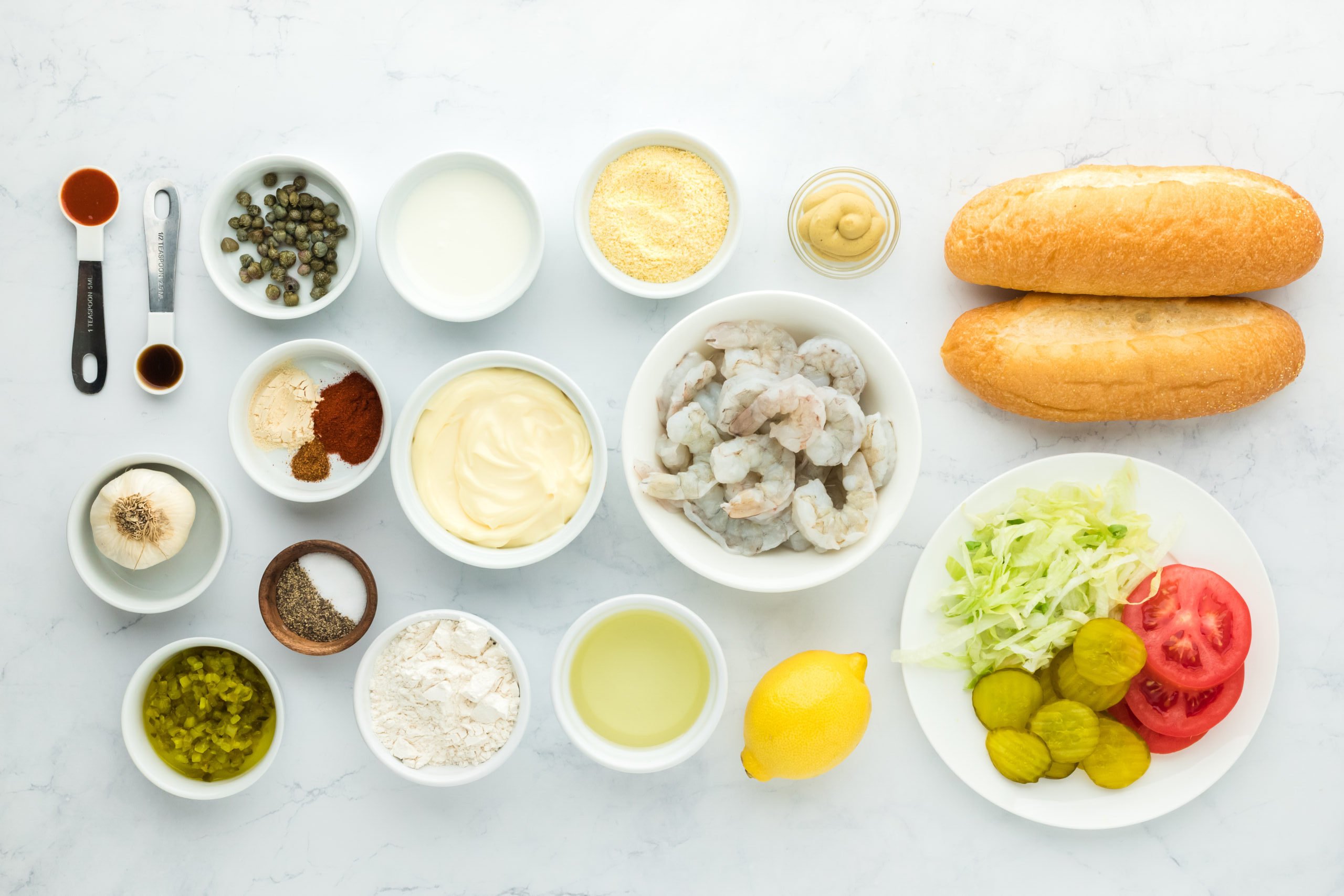 Bread, fresh shrimp, tomatoes and lettuce, spices, cornmeal and lemon juice in white bowls on white countertop