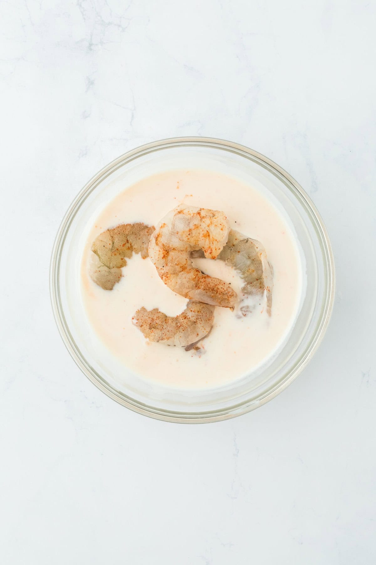 Raw shrimp added to a buttermilk mixture in a glass bowl on a white countertop