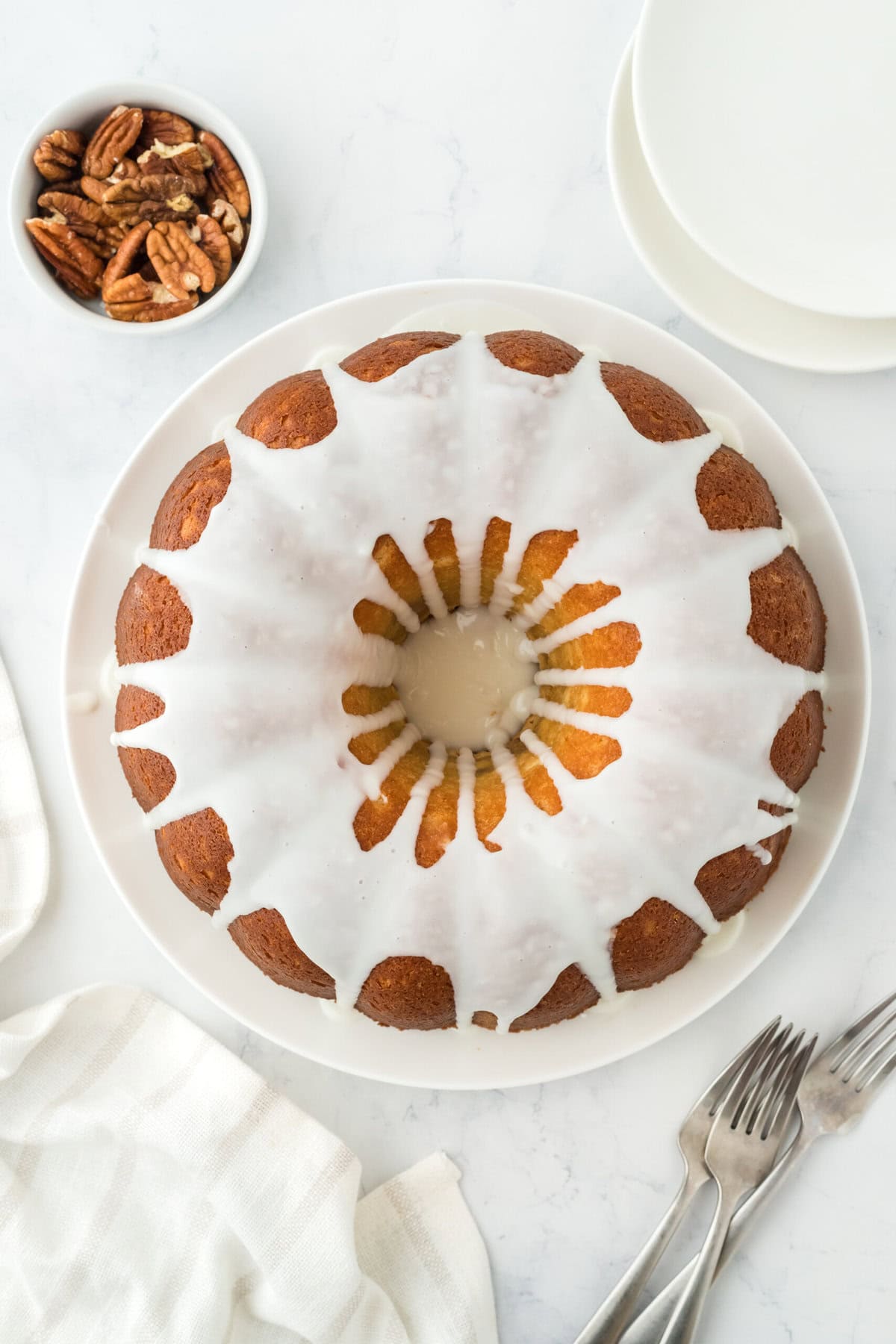 Homemade sock-it-to-me cake overhead with white icing on a white plate with pecans in a white bowl on white background ready to serve