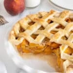 Fresh peach pie in a white dish with a slice removed, revealing the glistening peach filling and golden lattice crust, with milk bottles and plates in the background