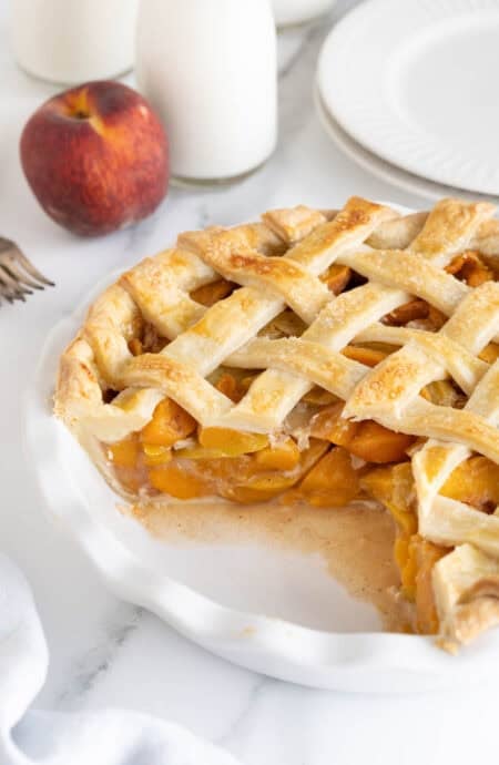 Fresh peach pie in a white dish with a slice removed, revealing the glistening peach filling and golden lattice crust, with milk bottles and plates in the background