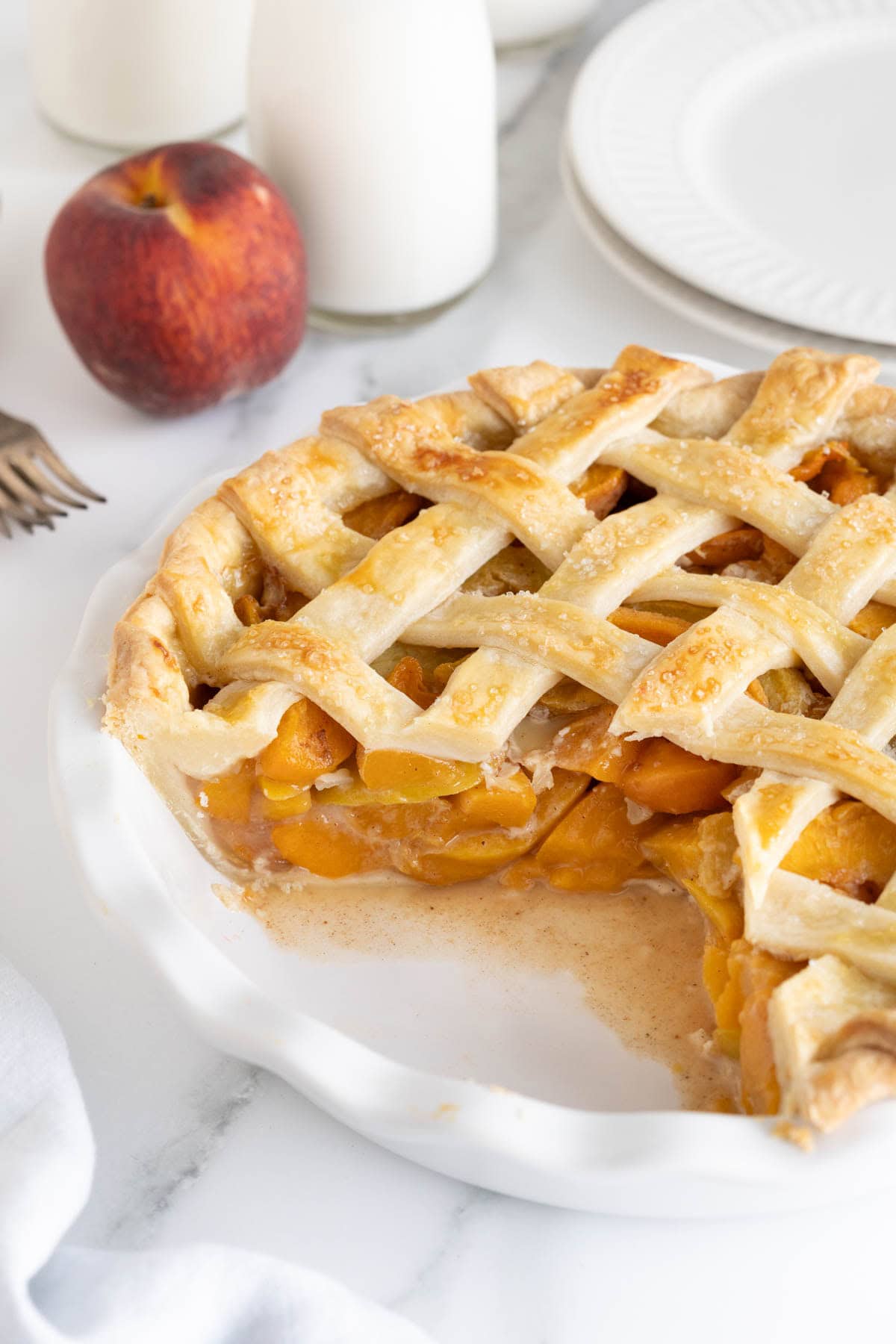 Fresh peach pie in a white dish with a slice removed, revealing the glistening peach filling and golden lattice crust, with milk bottles and plates in the background