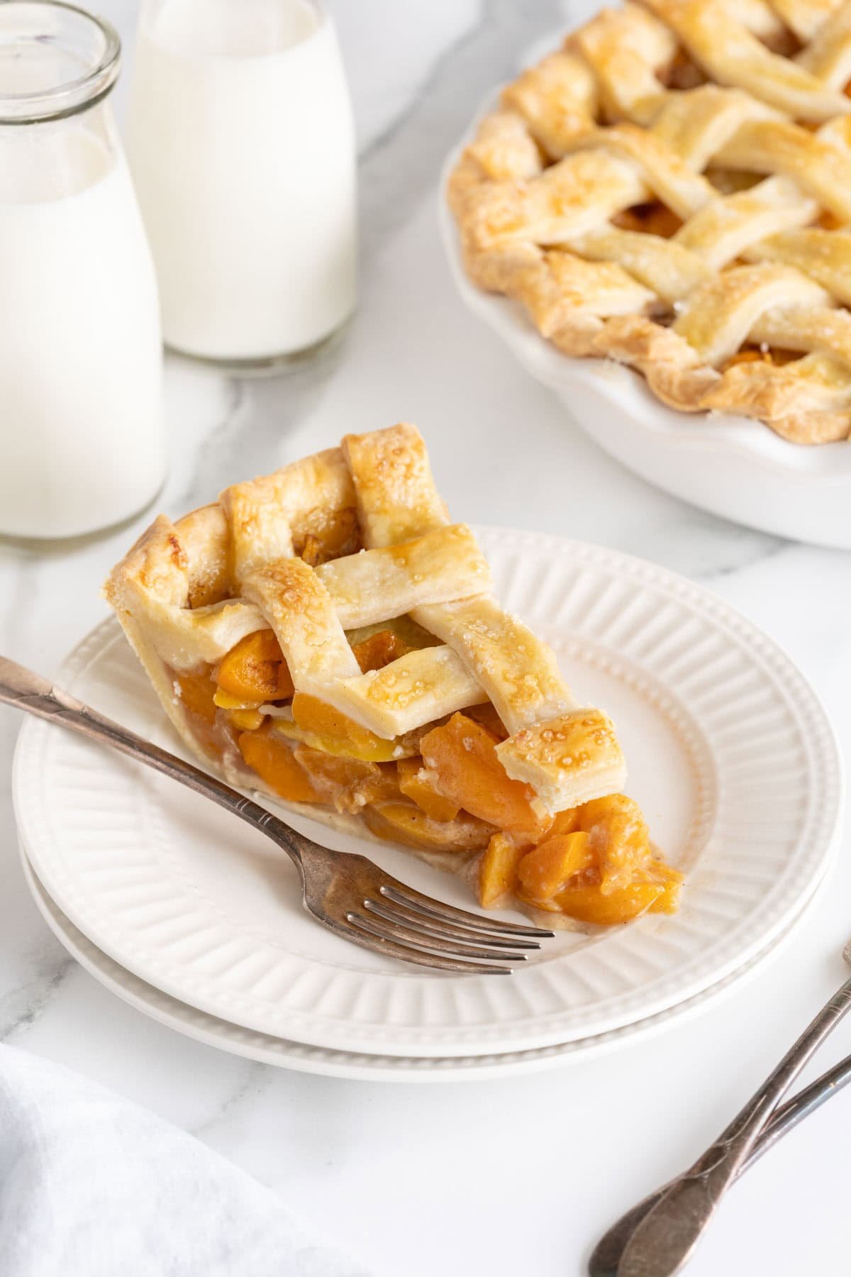 A slice of golden fresh peach pie with a lattice crust on a white plate, positioned next to a silver fork, with the rest of the pie and milk bottles blurred in the background