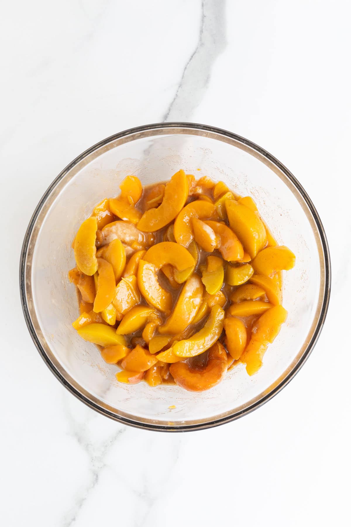 An overhead shot of a clear glass bowl filled with sliced peaches coated in a syrupy mixture, sitting on a white marble surface
