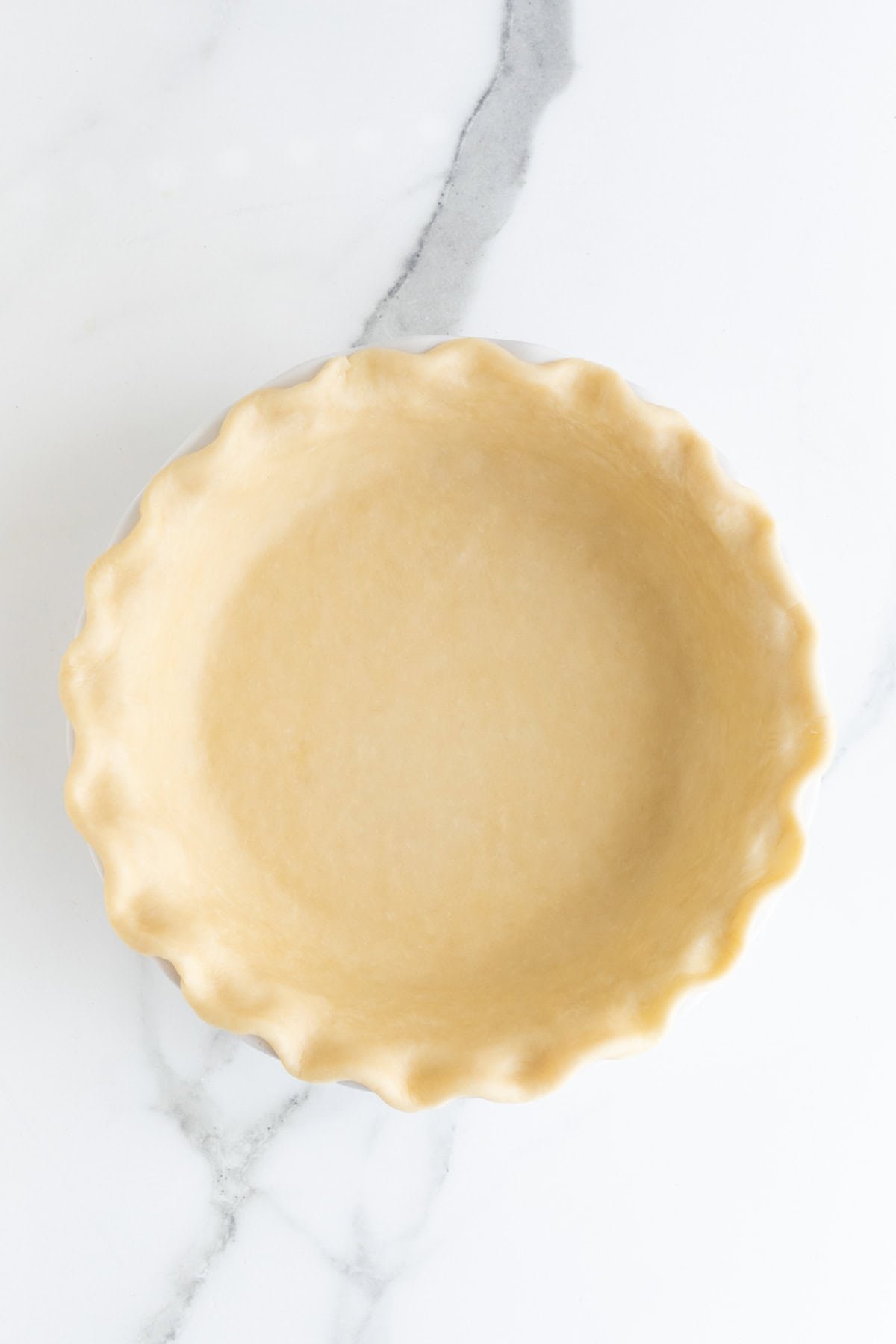 An overhead shot of an unbaked pie crust, perfectly crimped around the edges, sitting in a white pie dish on a white marble surface