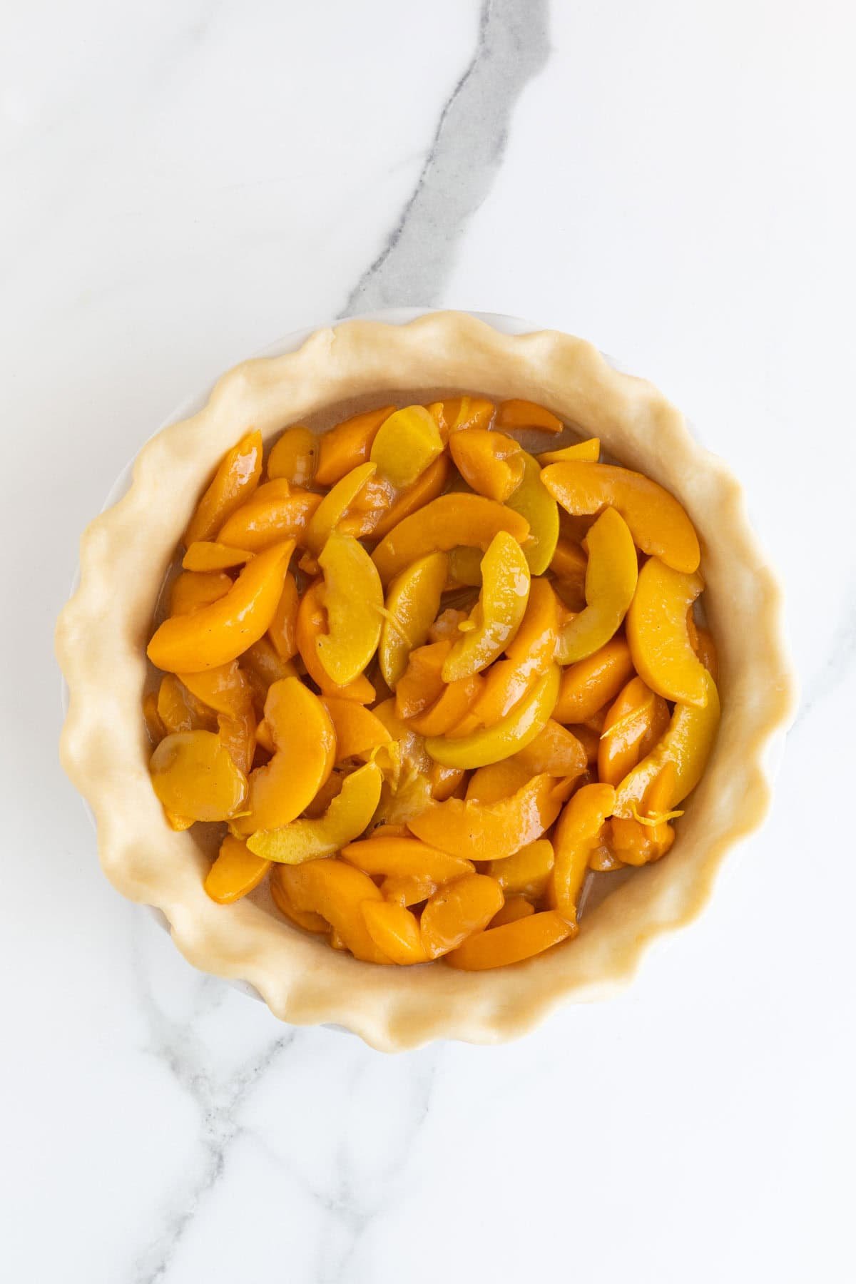 An overhead shot of an unbaked pie crust in a white dish, filled with a mixture of sliced peaches, on a white marble surface