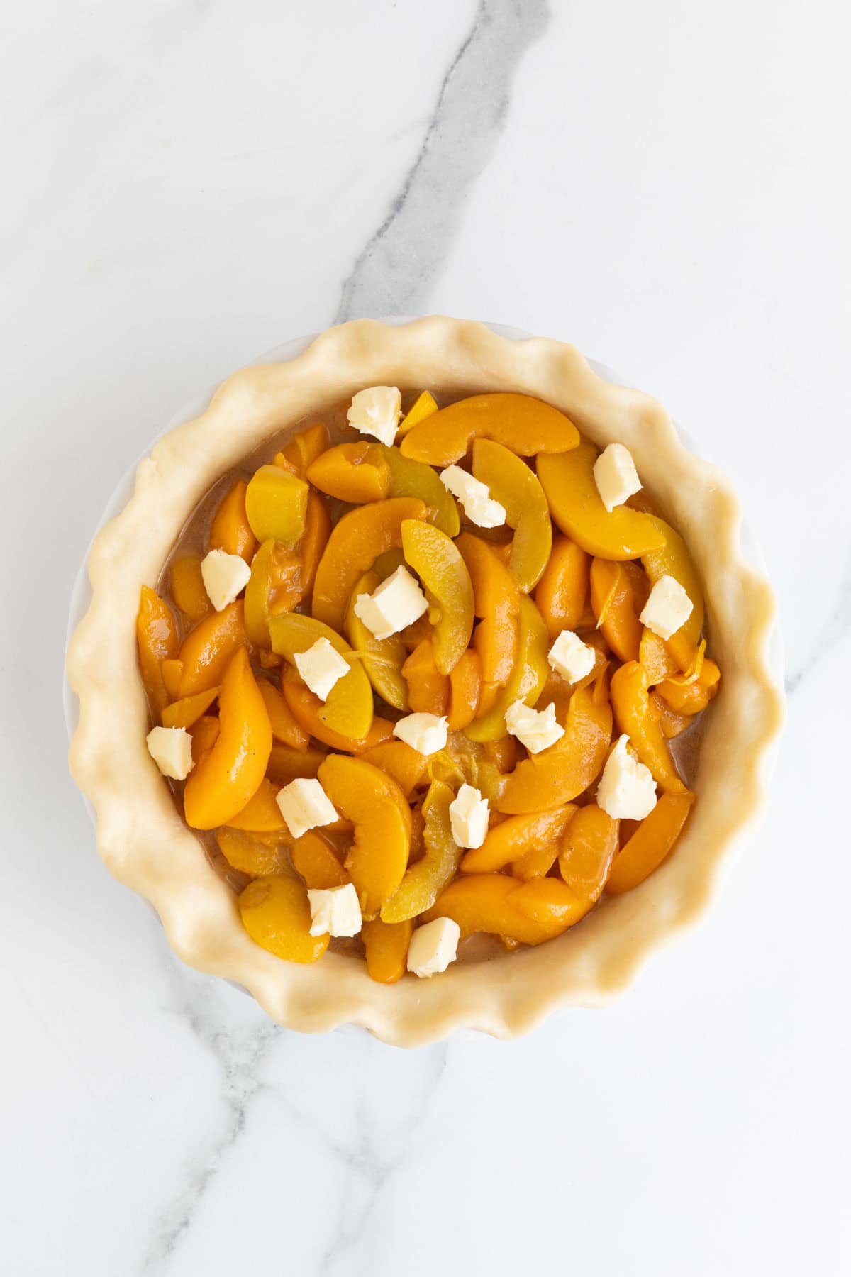 An overhead shot of a raw pie crust in a white dish, filled with sliced peaches and topped with small pats of butter, on a white marble surface