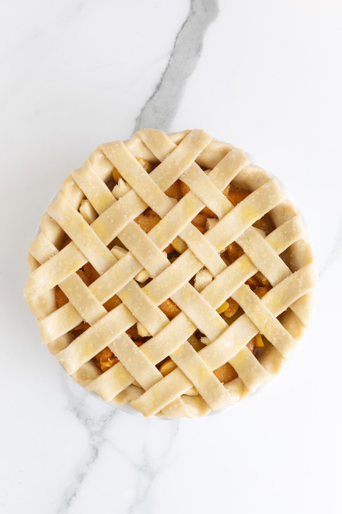 An overhead shot of an unbaked peach pie in a white dish, topped with a raw dough lattice crust, sitting on a white marble surface
