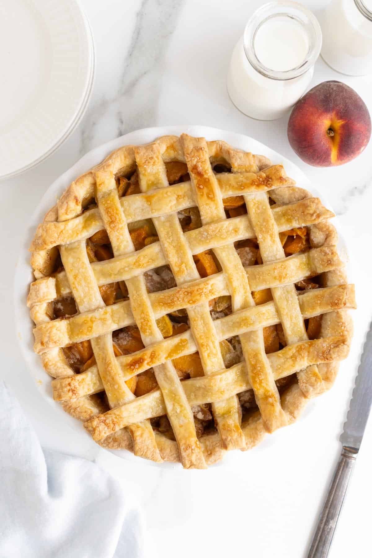 An overhead shot of a baked fresh peach pie with a golden-brown lattice crust in a white pie dish, alongside a fresh peach, milk bottles, and white plates on a marble surface