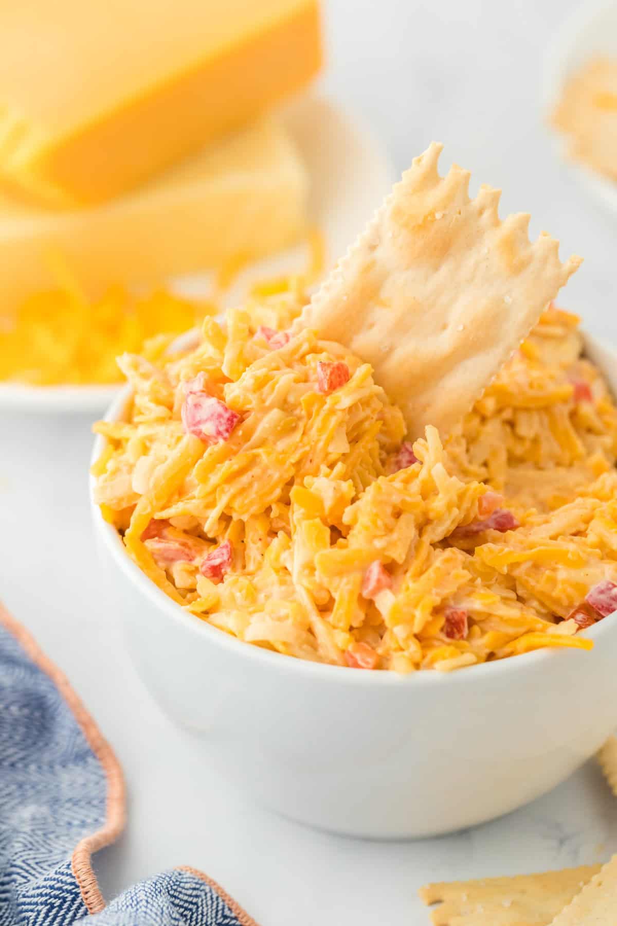A white bowl filled with Southern pimento cheese with a cracker inserted in the top and cheeses in the white background