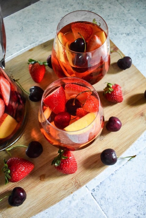 Two small glasses of Summer Fruit Rose Sangria surrounded by fresh strawberries and cherries on a wooden cutting board