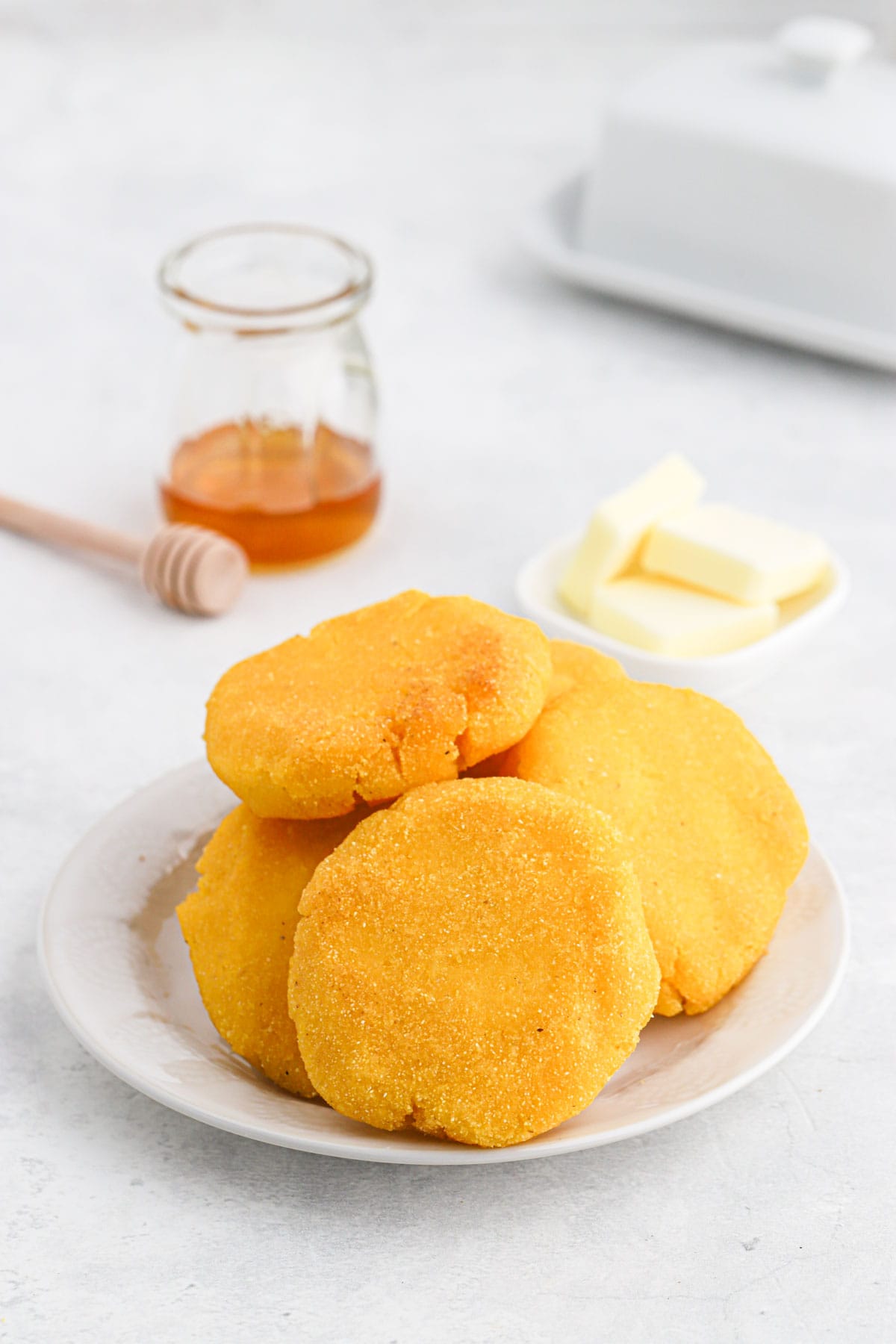 A pile of hot water cornbread on a white plate with a little jar of honey and a plate of butter.