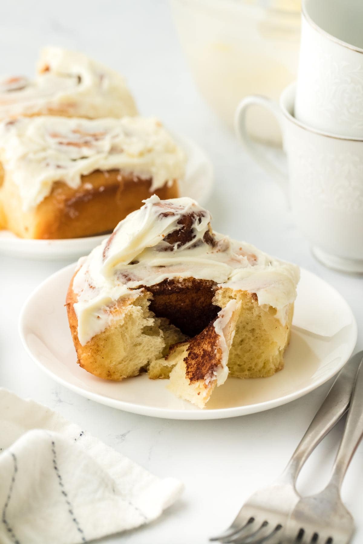 A closeup of a homemade cinnamon roll on a plate, topped with creamy frosting, with a bite taken out to reveal the soft, fluffy interior