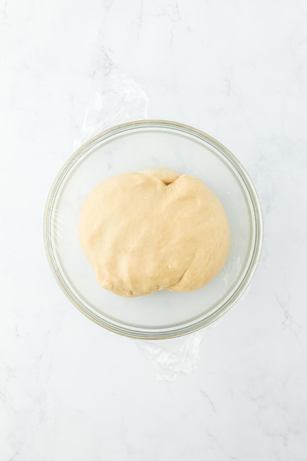 A glass bowl with a ball of smooth dough ready to rise, covered in plastic wrap