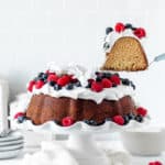 A slice of butter pound cake being lifted out of full cake with berries on white cake plate