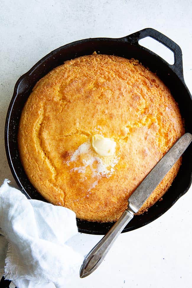 An overview photo of a large cast iron skillet filled with freshly baked traditional Cornbread recipe with butter melting over it and a knife for cutting