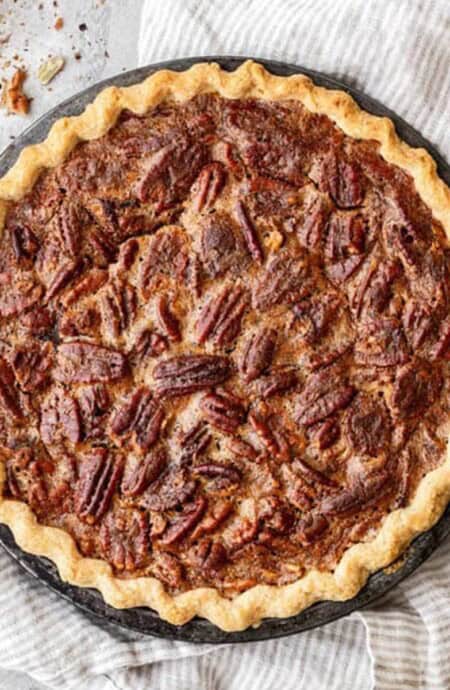 A whole southern pecan pie on the counter with pecans and a tea towel.