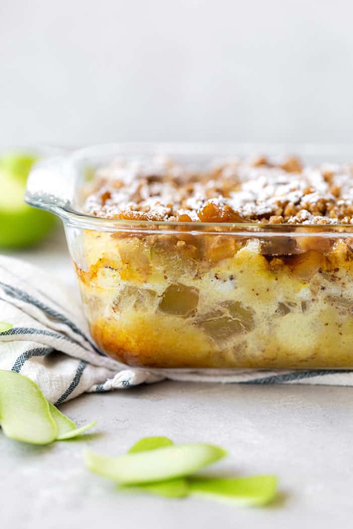 A powdered sugar sprinkled Apple Crisp French Toast Casserole ready to dig in with apples in background