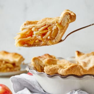 A slice of perfectly goldenbown double crusted apple pie recipe against gray background with a red apple being peeled