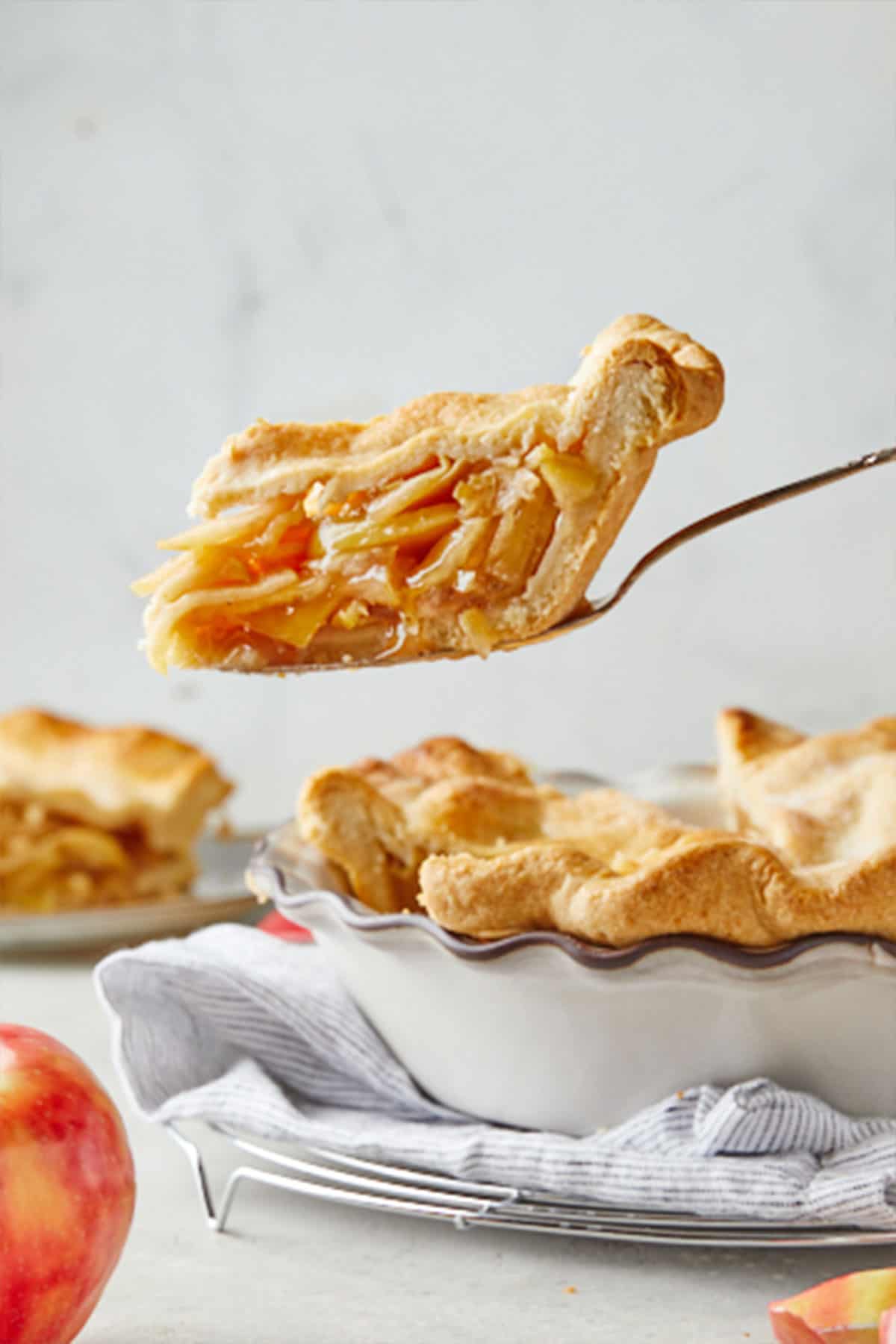 A slice of perfectly goldenbown double crusted apple pie recipe against gray background with a red apple being peeled