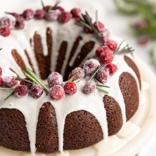 A gingerbread cake on a cake stand topped with glaze, cranberries, and fresh rosemary.