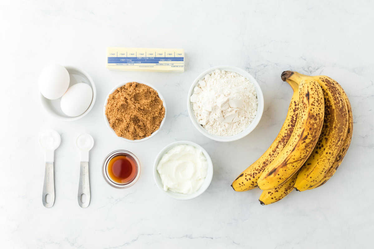 Ripe bananas, flour, leavening, brown sugar, eggs, sour cream, vanilla and butter on a white background