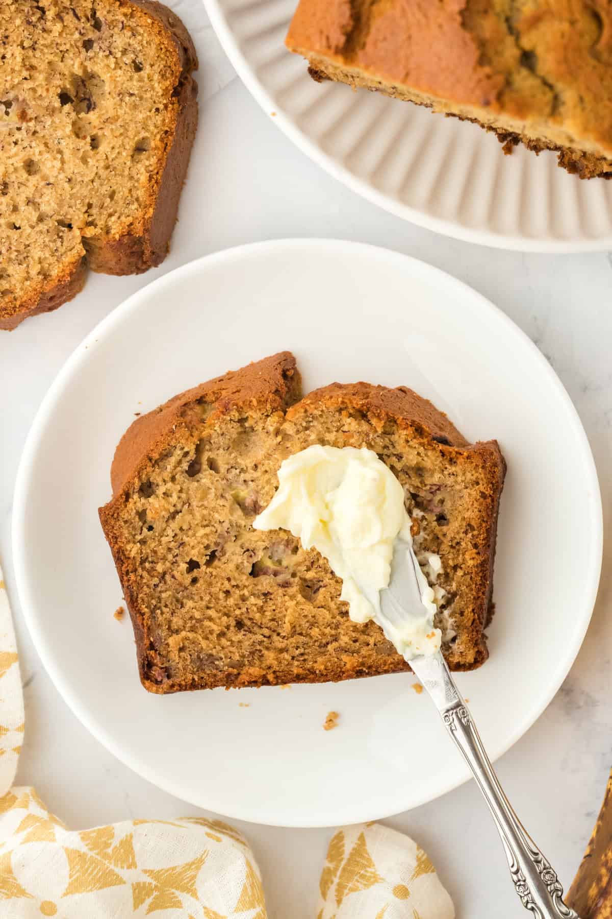 Homemade banana bread slice on a white plate with butter being smeared on top of it with a white and yellow napkin in the white background