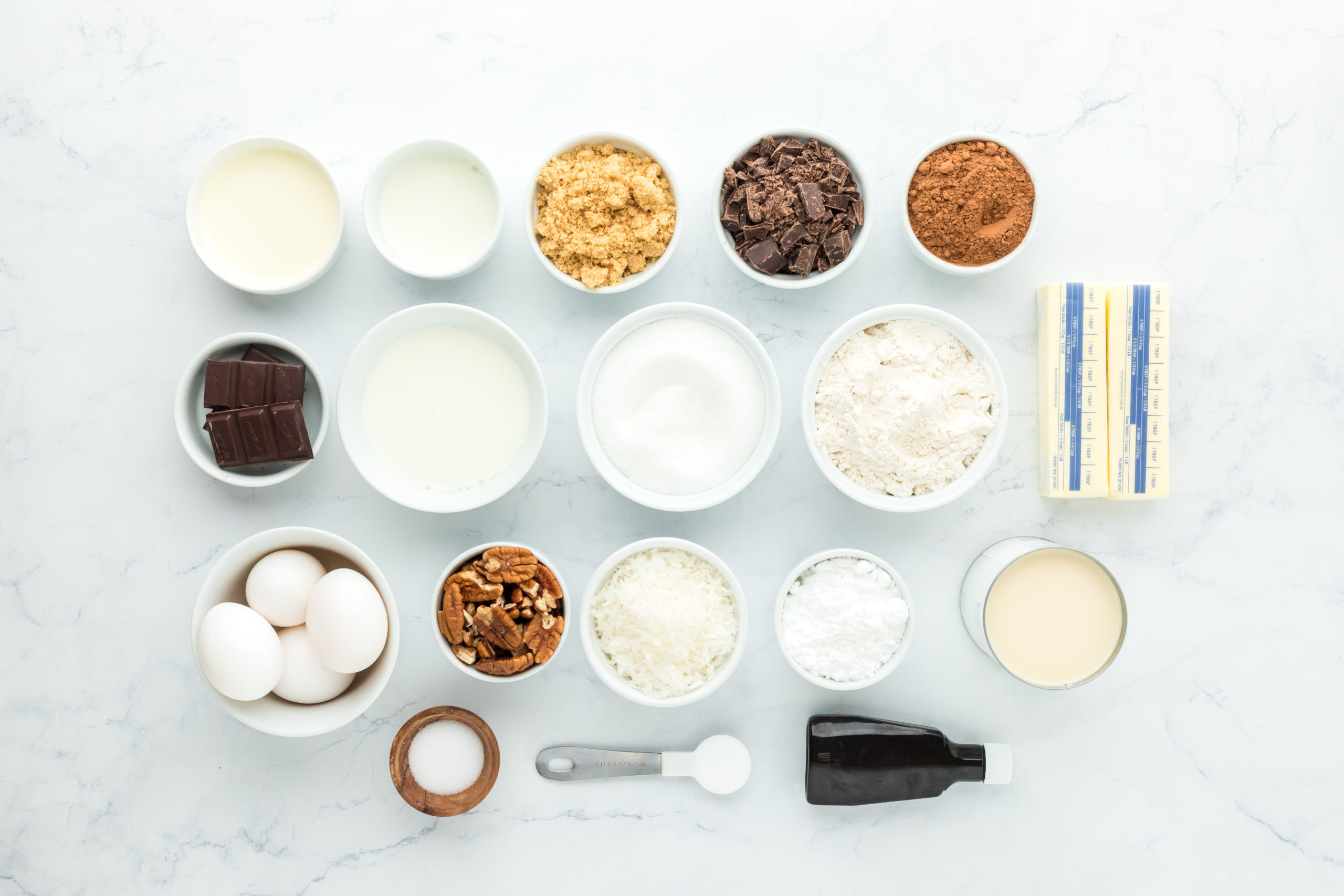 Several small white bowls with butter, sugar, chocolate, eggs and vanilla on white countertop