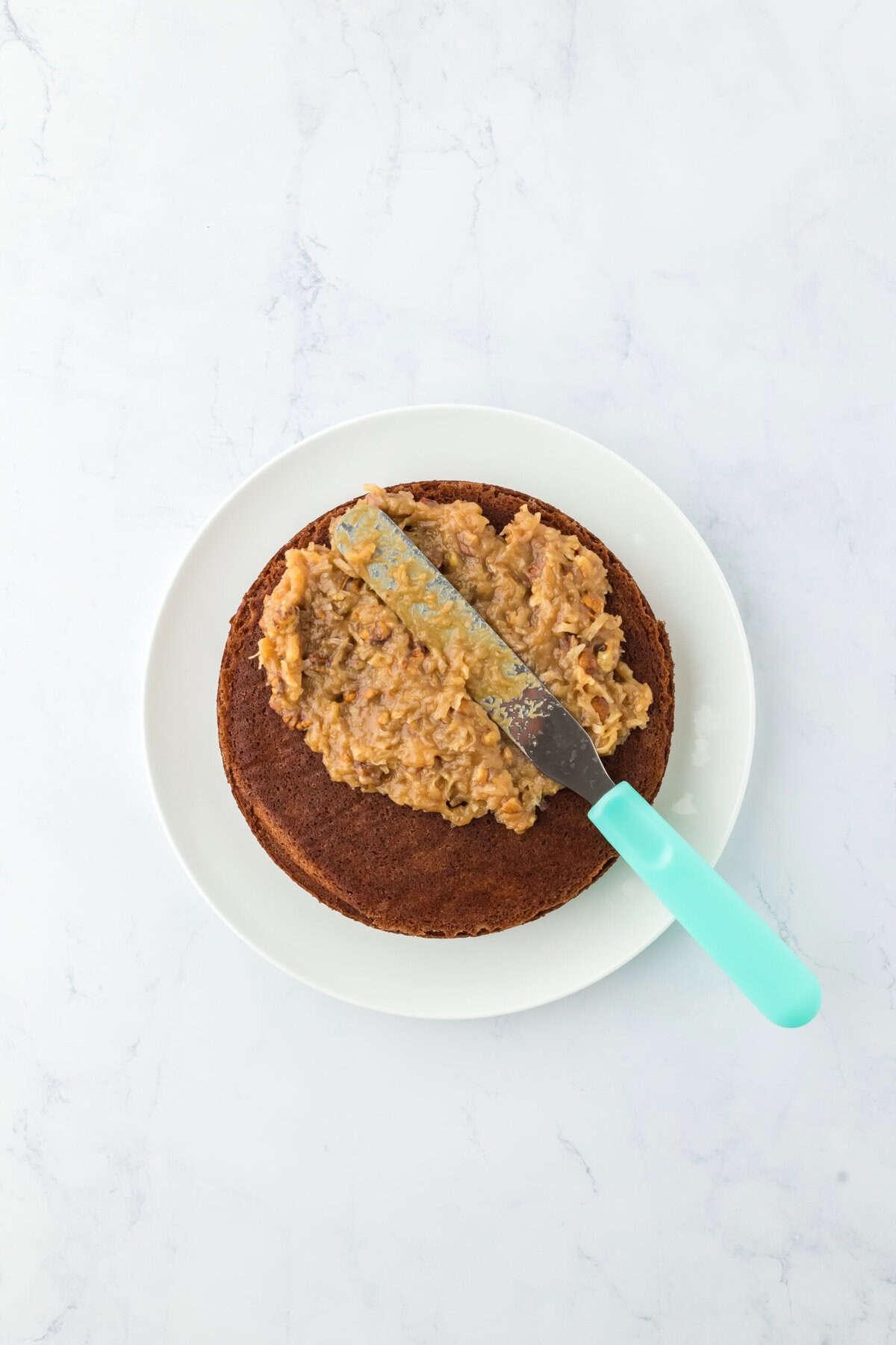 German chocolate cake icing being spread on cooled layer of cake on white countertop.