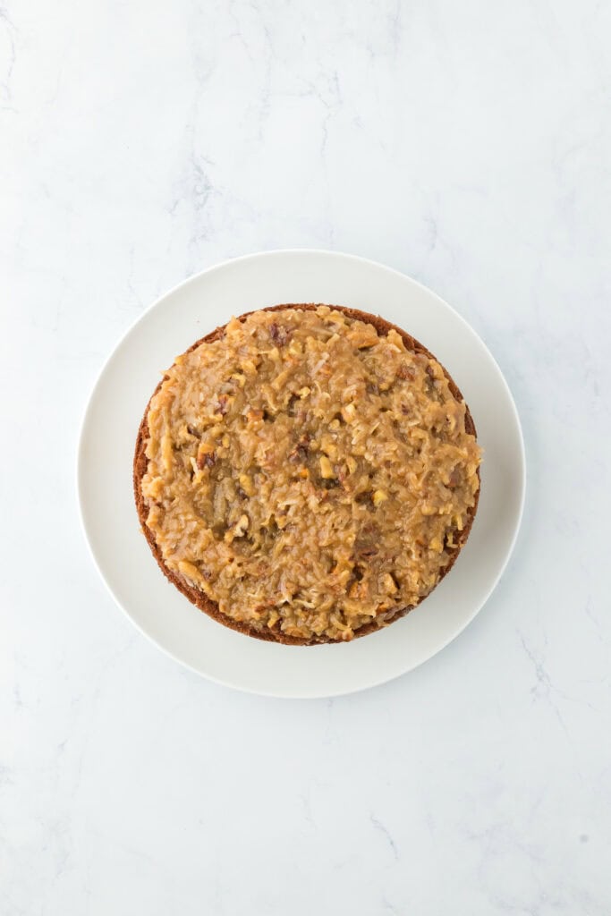 Coconut and pecan Icing added to a layer of chocolate cake on a white countertop