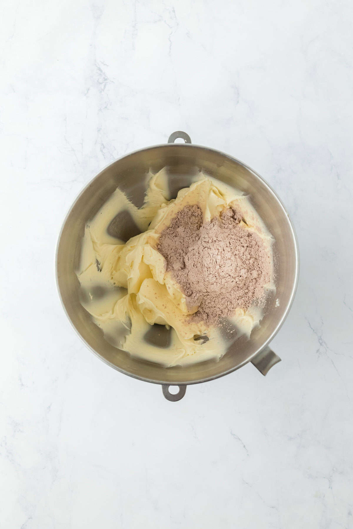 Butter, confectioner's sugar and chocolate in a stand mixer bowl on white countertop.