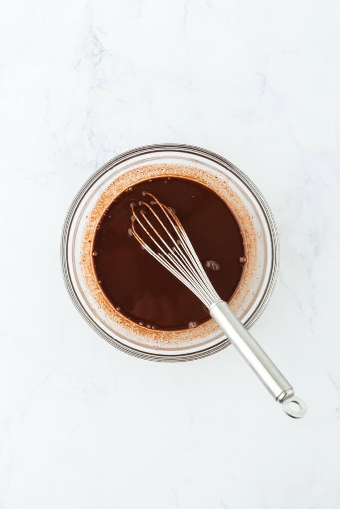 Chocolate sauce with a whisk in glass bowl on white countertop.