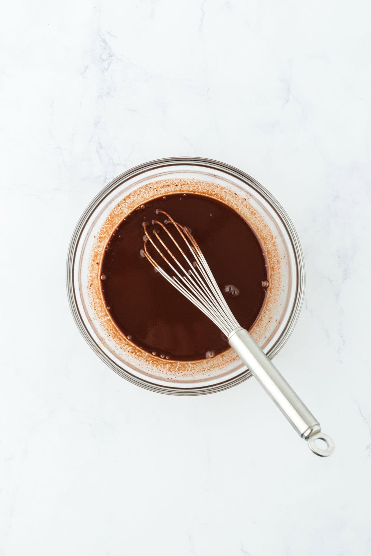 Chocolate sauce with a whisk in glass bowl on white countertop.