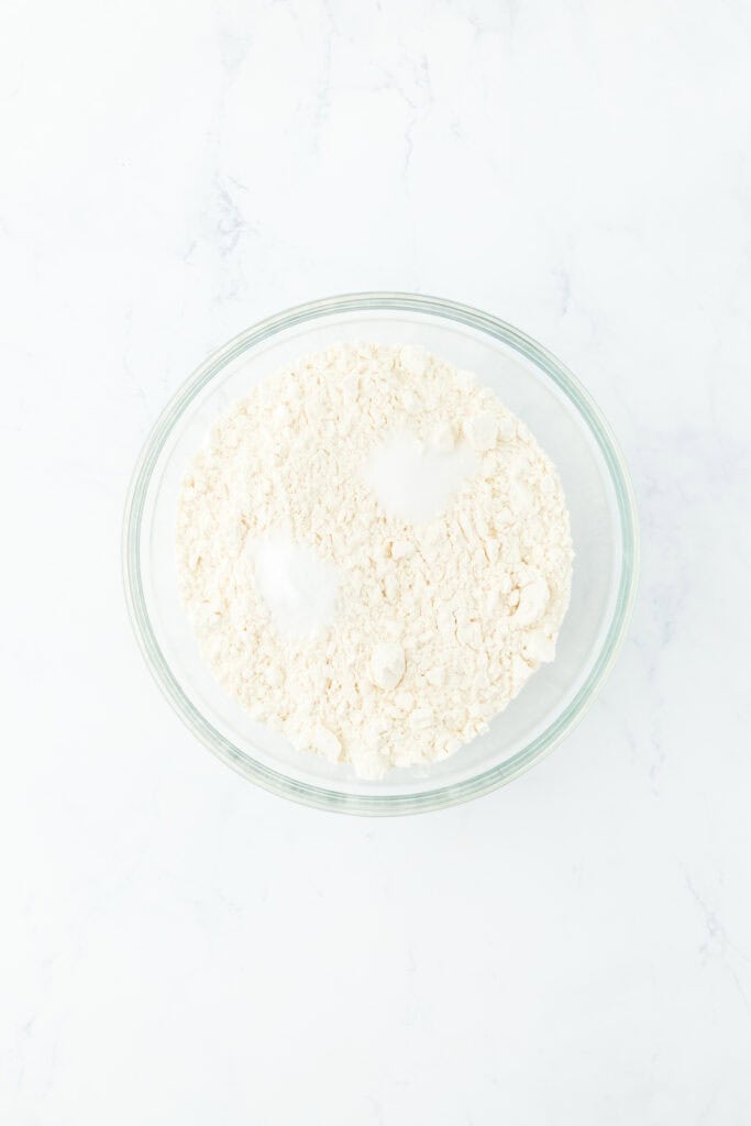 Dry ingredients being added to glass bowl on white countertop.