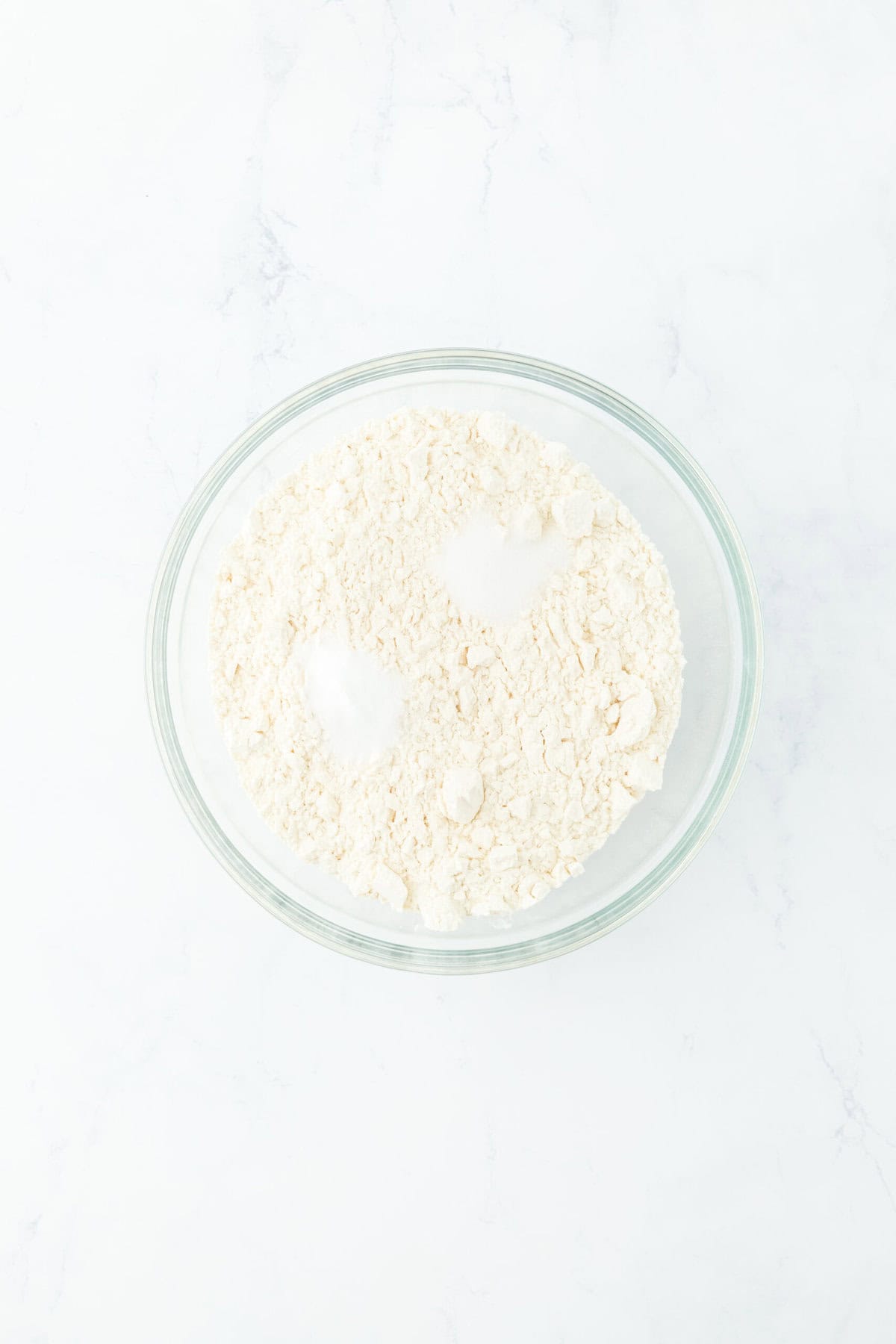 Dry ingredients being added to glass bowl on white countertop.