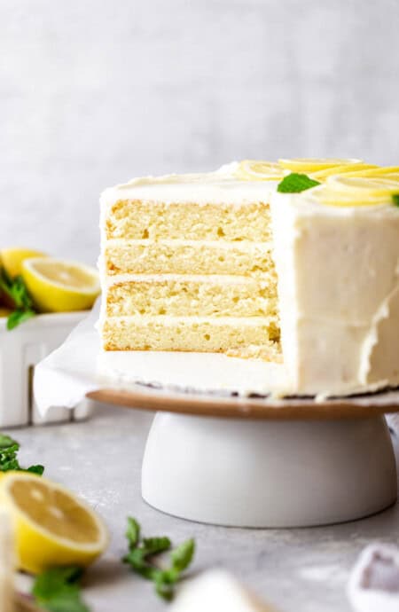 A delicious lemon layer cake sliced into with lemons in the background on a white cake stand
