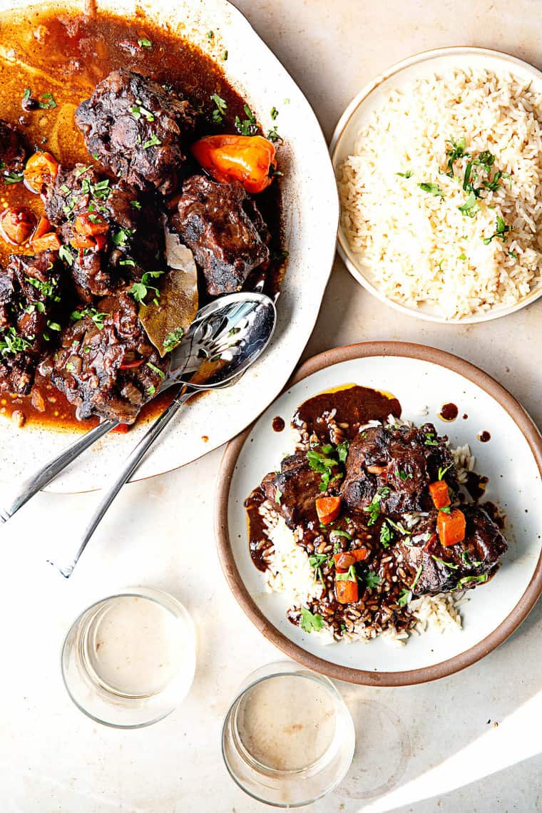 Delicious oxtails served over rice in a white plate with a platter of Jamaican oxtails in the background