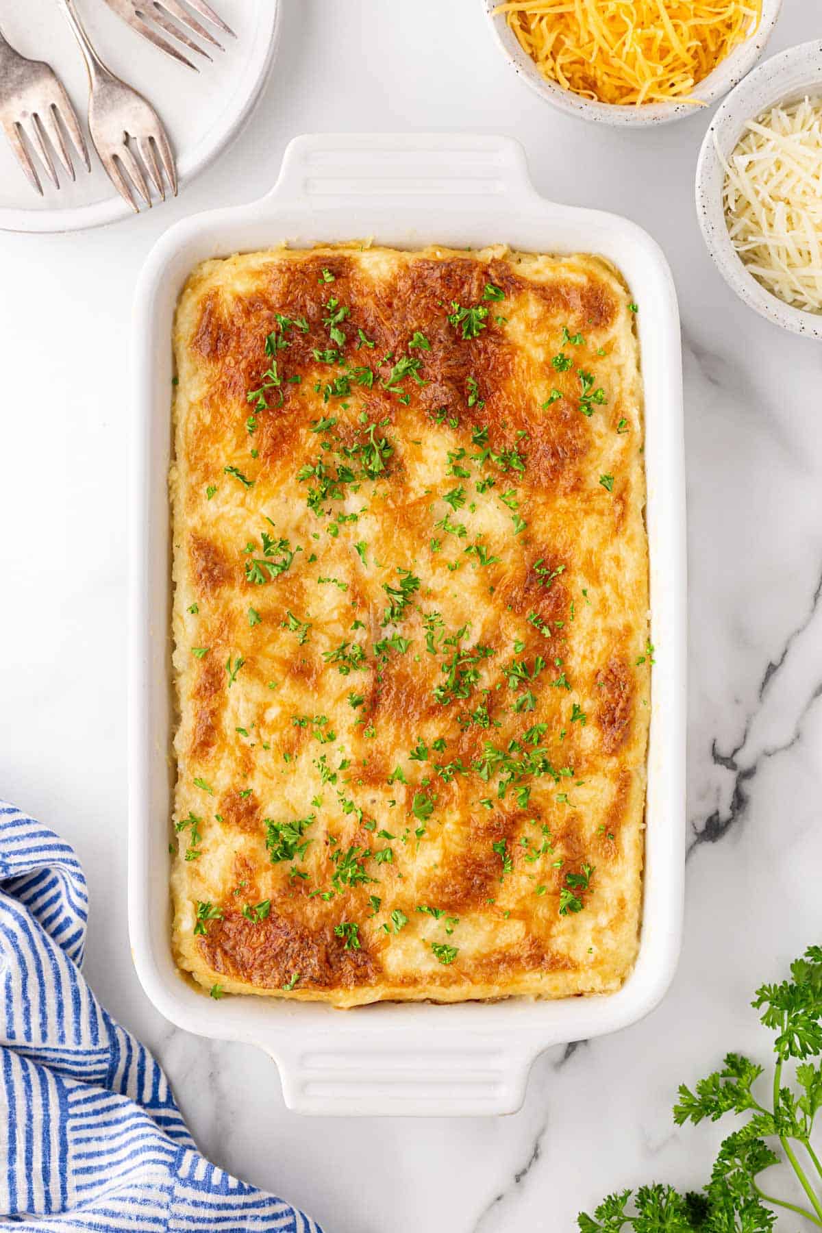 Golden baked grits casserole topped with parsley, with shredded cheeses, silverware, and a striped towel nearby