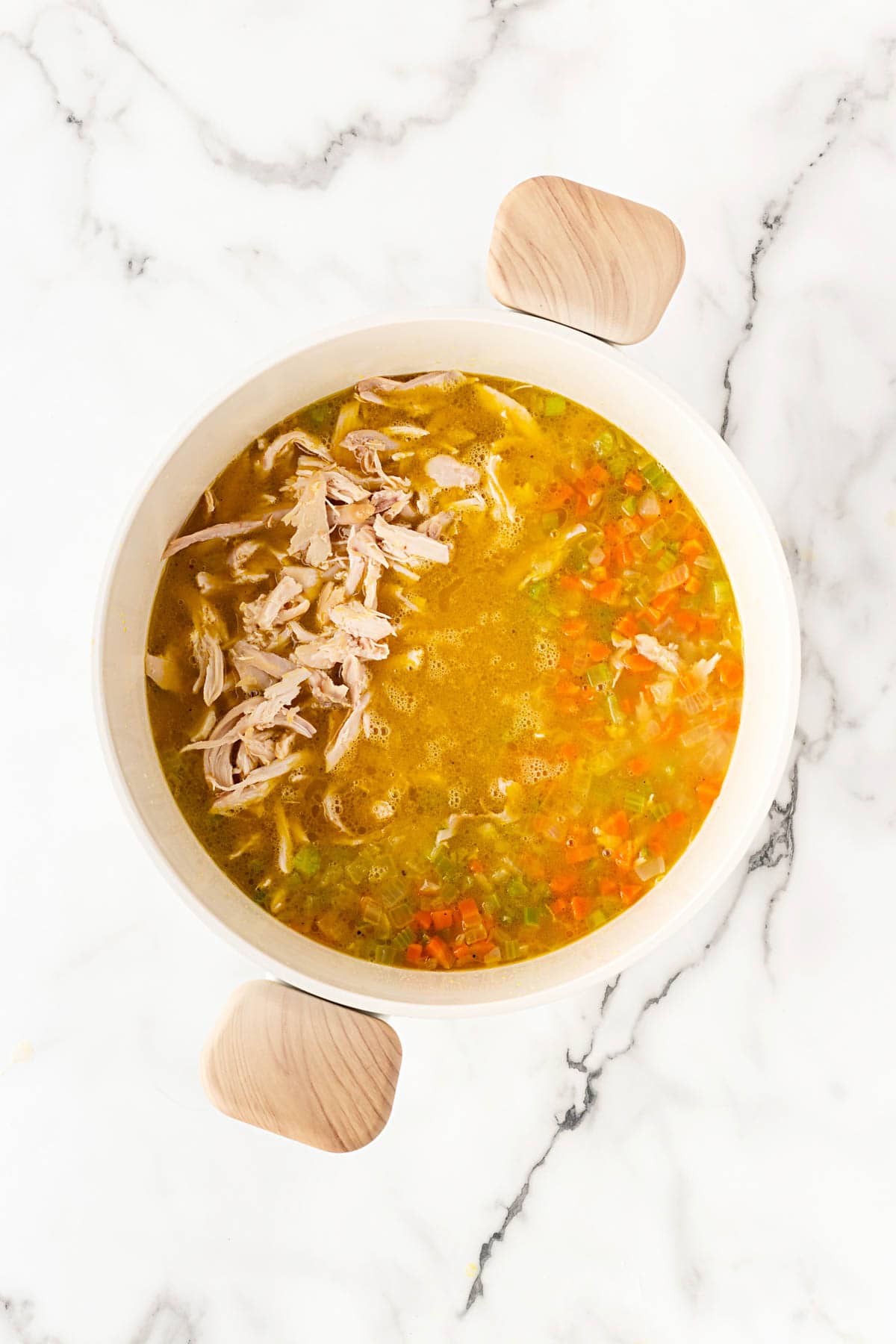 Overhead shot of shredded chicken added to a pot of simmering broth with diced carrots, celery, and onions