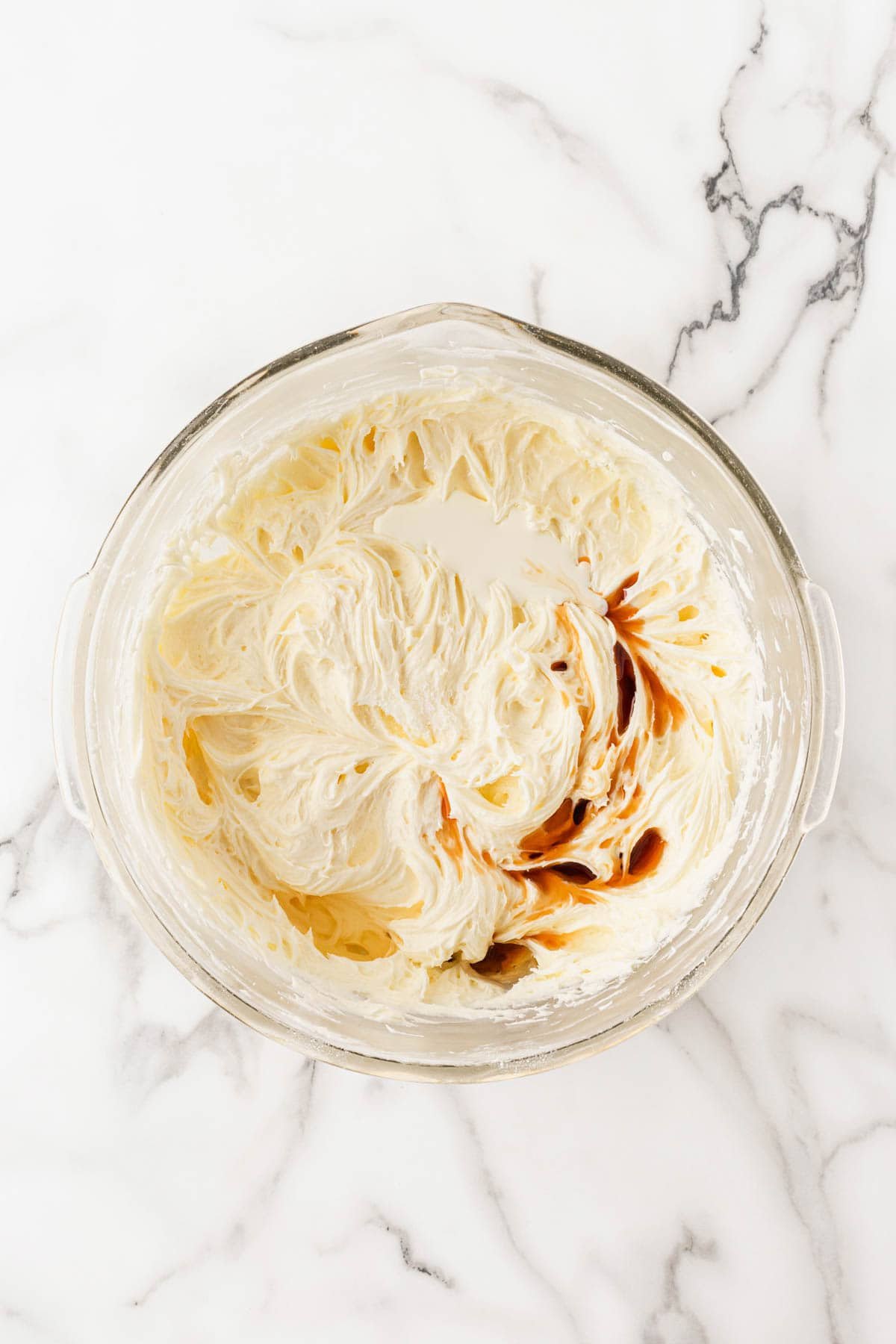 A mixing bowl of lemon cream cheese frosting being blended with heavy cream, vanilla, lemon extract and salt