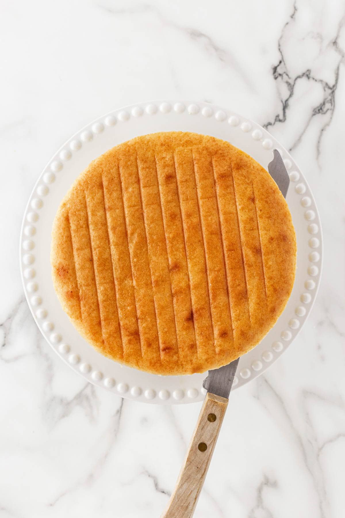 A baked cake layer on a plate with lined impressions on top and a serrated knife cutting it