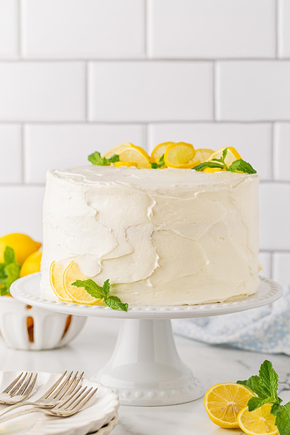 A whole lemon layer cake on a cake stand, frosted in white and topped with lemon slices and mint