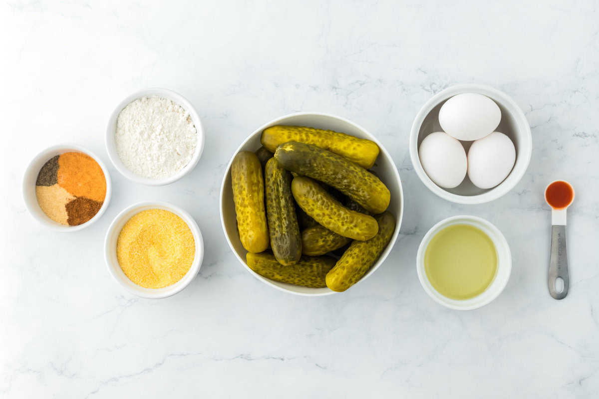 Pickles, flour, eggs, seasonings and cornmeal in white bowls against a white background