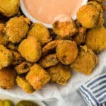 Fried pickles on a white bowl with a dipping sauce and dill pickles in the background