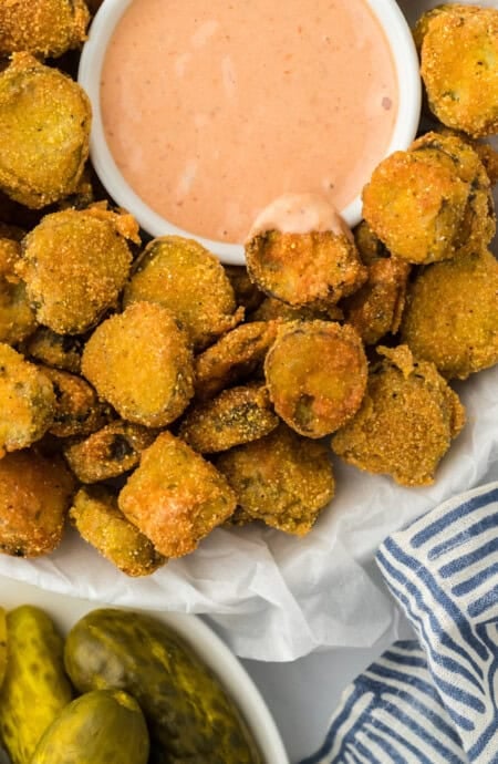 Fried pickles on a white bowl with a dipping sauce and dill pickles in the background
