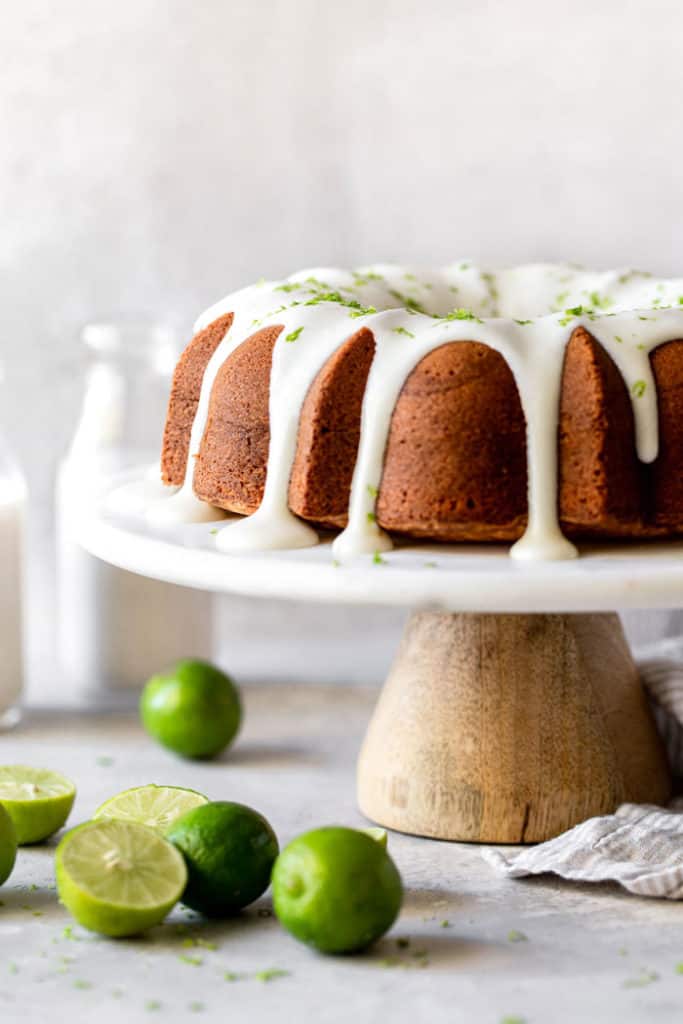 A key lime cake on white platter with key limes surrounding it ready to cut into