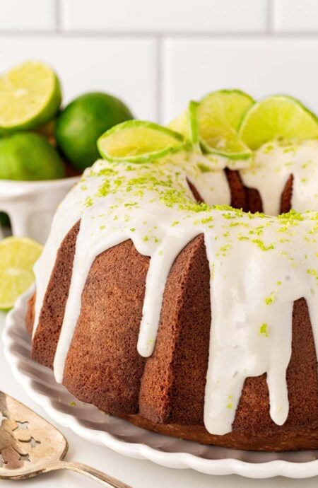Closeup of a key lime cake topped with white glaze, lime zest, and sliced limes on a serving platter