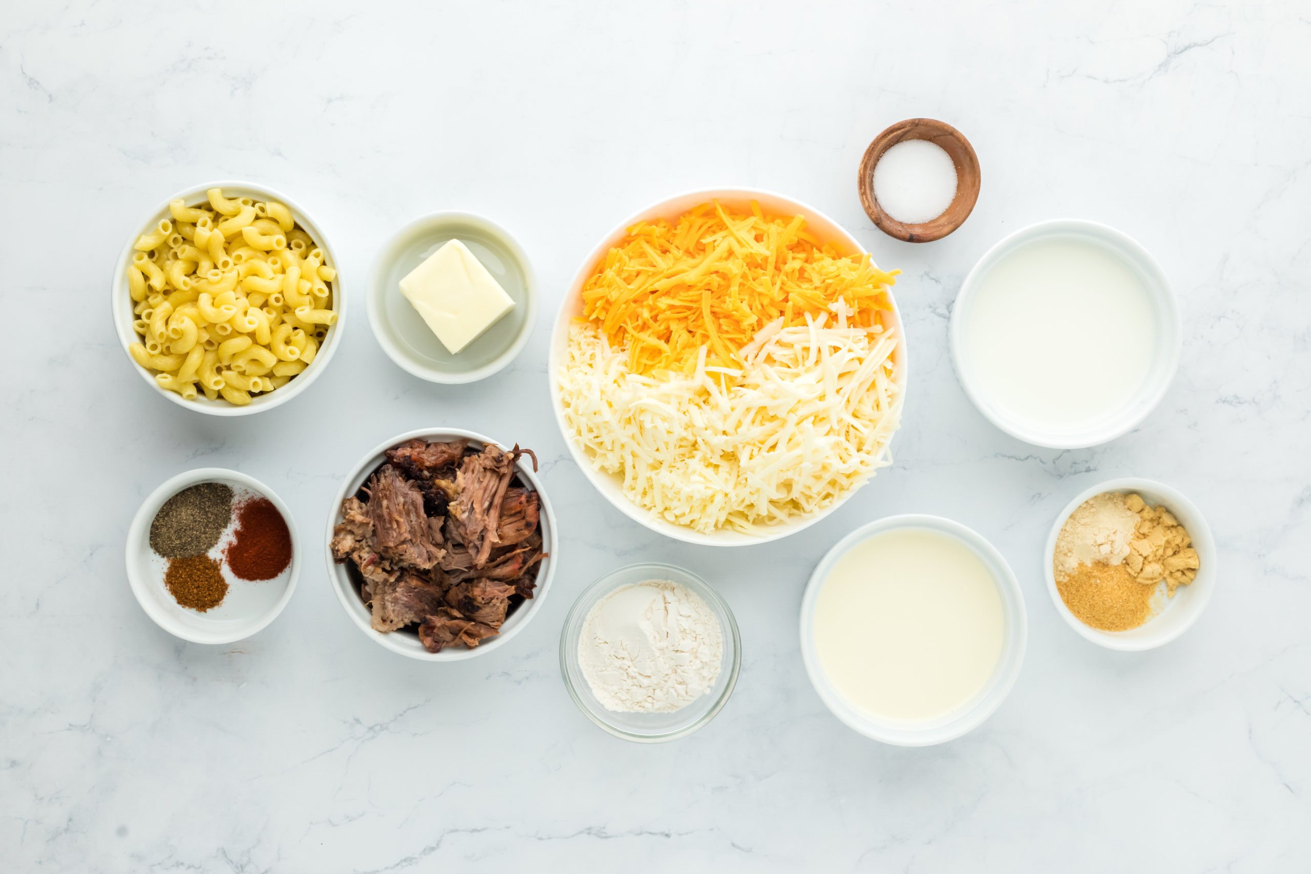 Brisket, shredded cheeses, milk, macaroni pasta, butter, seasonings in bowls on white countertop