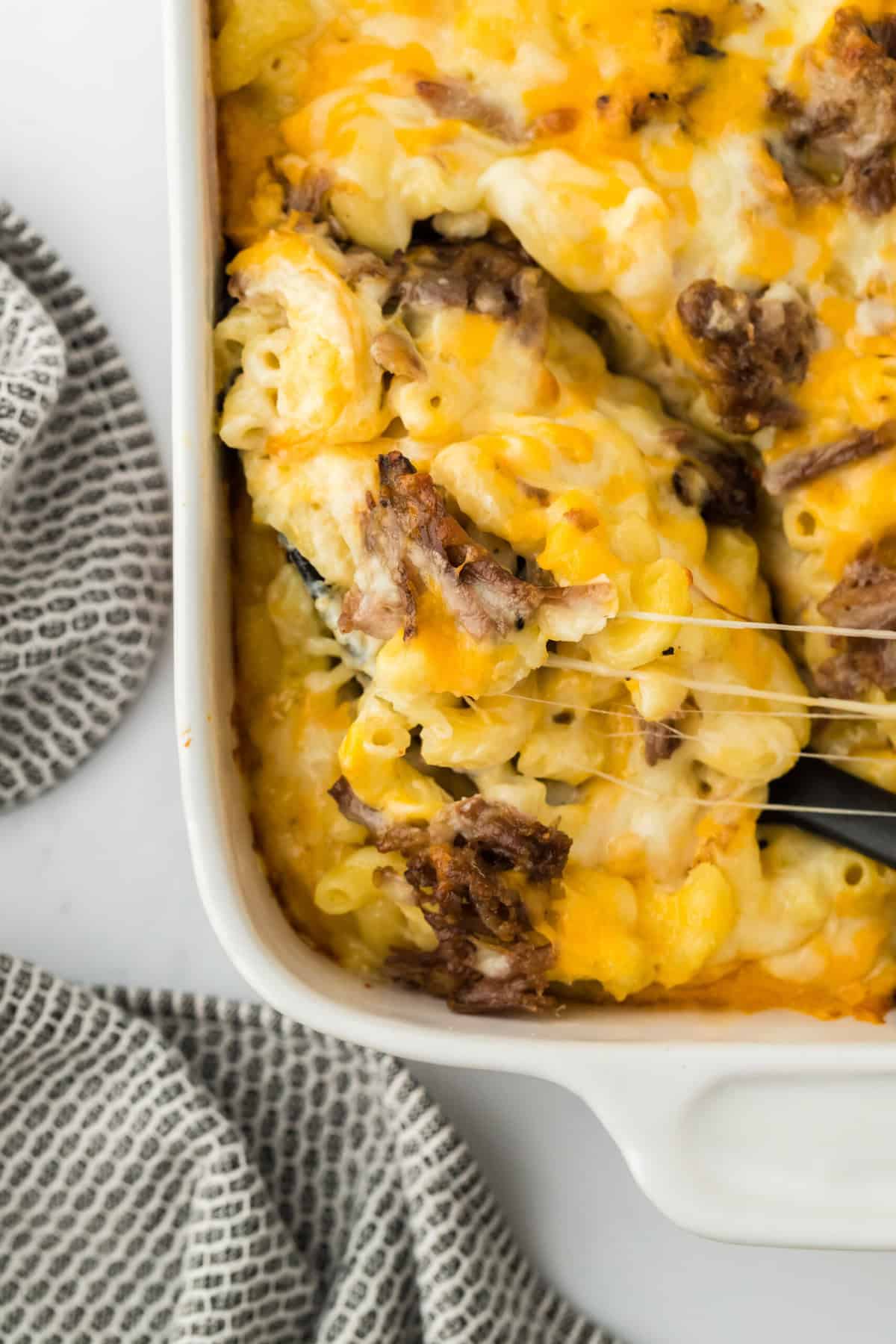 Brisket macaroni and cheese being lifted from a casserole dish to serve on white countertop