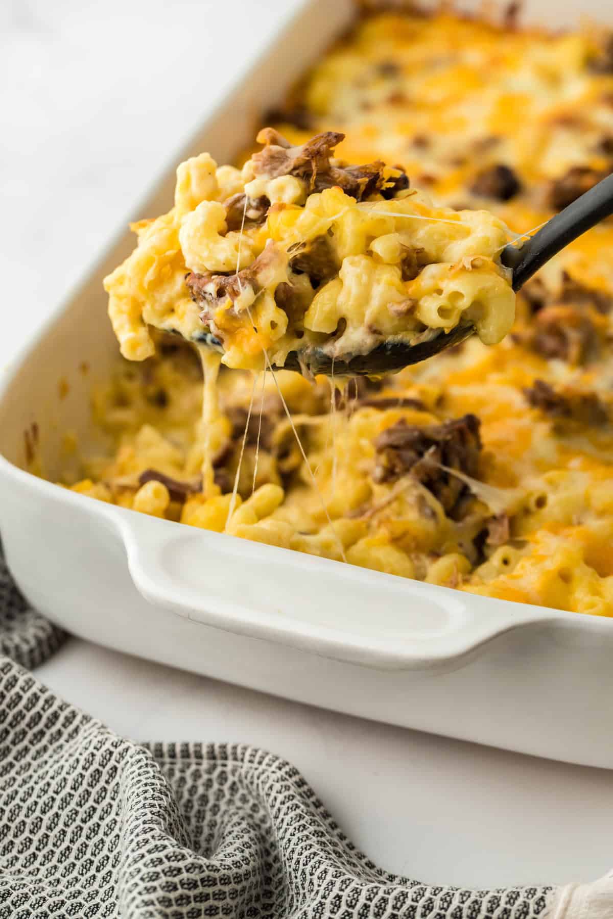 Beef brisket mac and cheese being lifted from a white casserole dish to serve